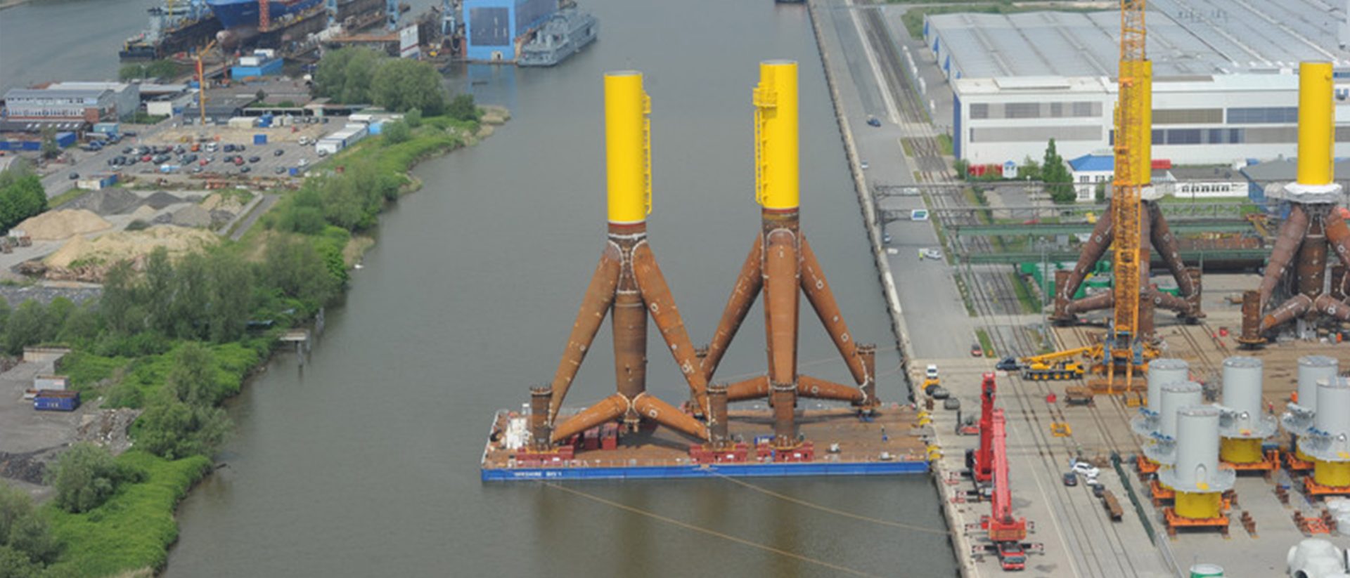 Aerial view of large offshore wind turbine foundation structures on a barge in an industrial harbor area with cranes and warehouses nearby.