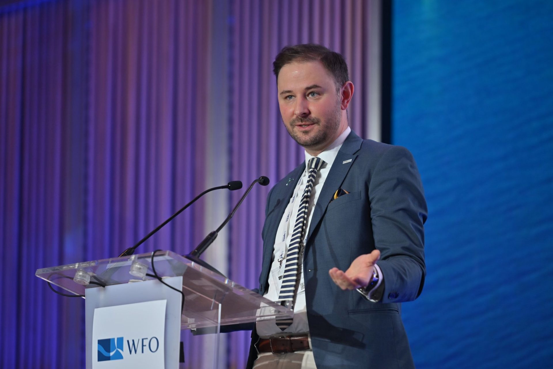 Ein Mann im blauen Anzug spricht während des WFO Global Summit 2025 an einem durchsichtigen Podium mit zwei Mikrofonen. Der Hintergrund ist blau beleuchtet und auf dem Podium befindet sich ein WFO-Logo.