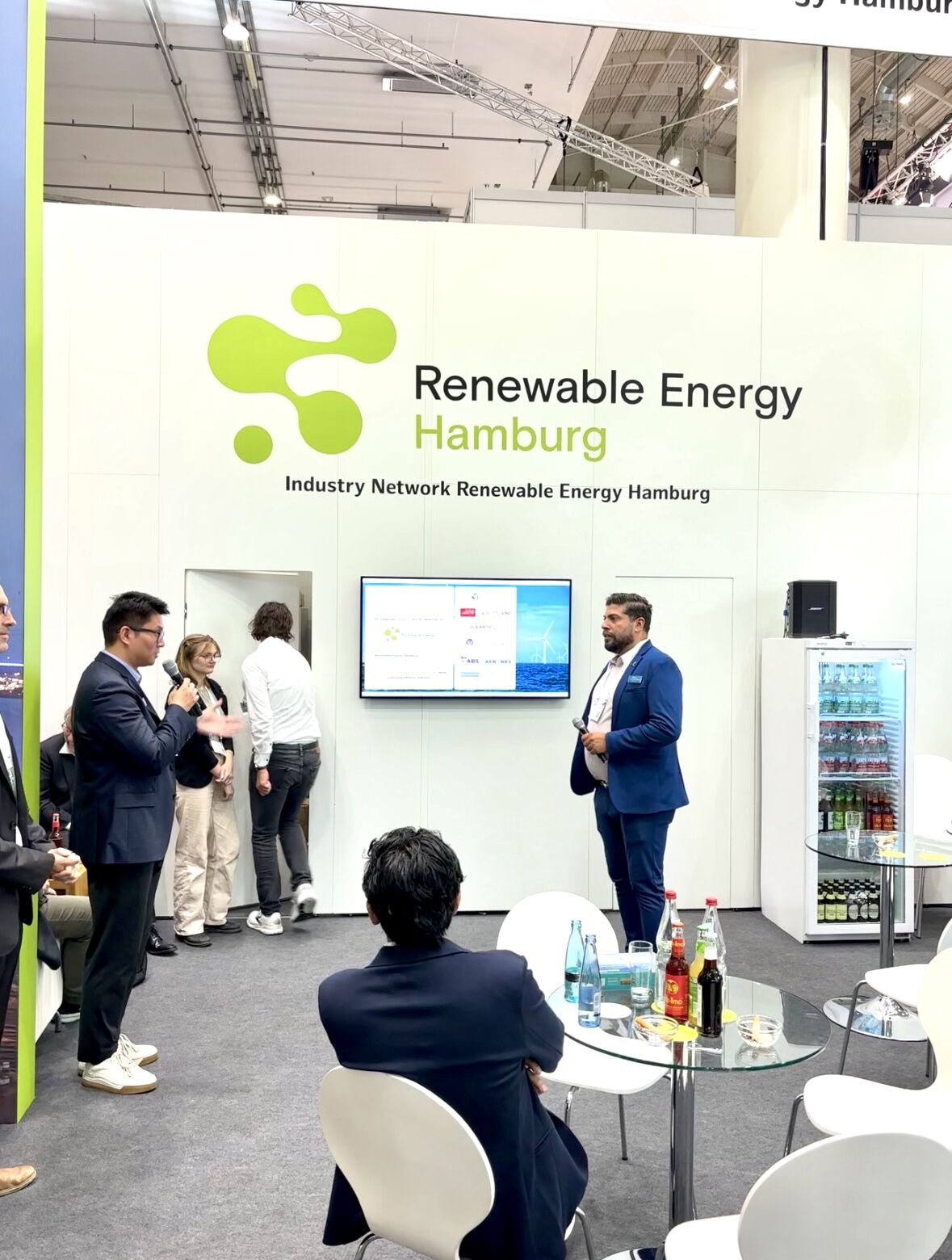 A group of people converse and present in a booth labeled "Renewable Energy Hamburg" at the WindEnergy Hamburg trade show, with a display screen and a fridge stocked with drinks.