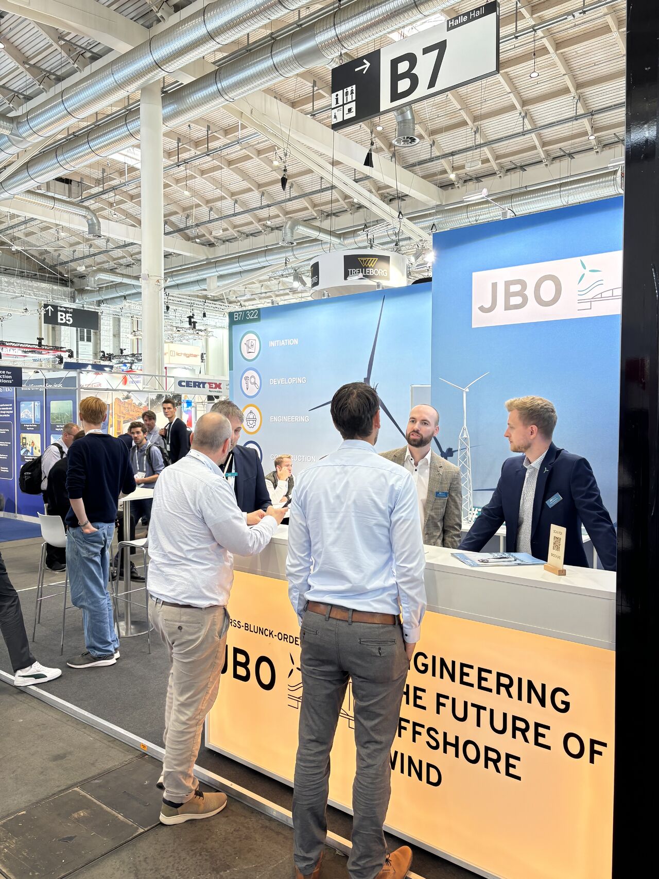 People interact at the JBO booth at WindEnergy Hamburg, with signage reading "Engineering the Future of Offshore Wind" and displays featuring wind turbines.