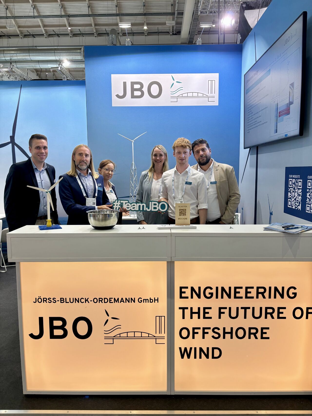 Six people stand behind a booth for JBO at WindEnergy Hamburg Day 1, featuring wind turbine models and informational displays promoting offshore wind engineering.